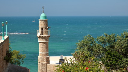 Krajobraz nadmorski z minaretem ozdobionym półksięzycem, taras widokowy, dzrewo, w tle szmaragodwe morze z niewielkimi falami,  jachty na wodzie, słonecznie