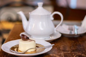 Florence, Tuscany / Italy. - April 21, 2018. Italian dessert: round vanilla cheesecake decorated with milk chocolate, served on a white plate, next to a white teapot on a wooden table 