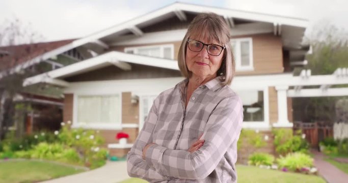 Confident senior woman with arms crossed standing outside suburban home, Mature old lady looking proud crossing arms outside nice house, 4k