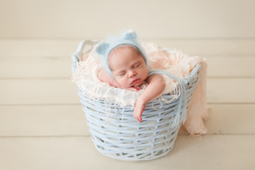 Newborn sleeping in a wicker basket in a blue fluffy knit kitty hat on a pink props on a beige...