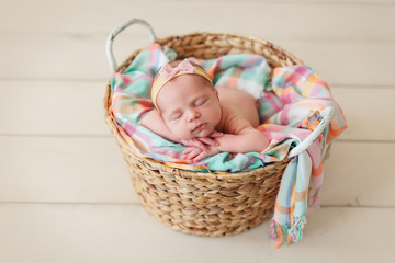 Newborn baby girl in bandage bow sleeping in wicker basket on plaid shawl on beige wooden floor