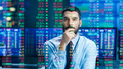 Stock Market Top Trader Looks at Projected Ticker Numbers and Graphs Running, Analysing Data to Make Best Sell. Behind Him Room Full of Screens and Statistics.