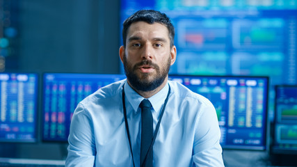Professional Stock Market Trader Charismatically Talks into the Camera. Behind Him Computer Screens with Ticker Numbers, Data, Graphs.