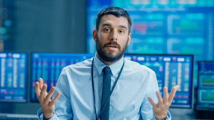 Professional Stock Market Trader Charismatically Talks into the Camera. Behind Him Computer Screens with Ticker Numbers, Data, Graphs.