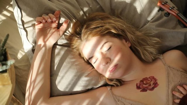 Overhead view of young blond girl with tattoo on chest sleeping on bed in rays of sunlight.