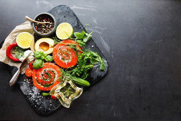 Delicious Salmon rolls, goats cheese in paper with knife, aromatic herbs, spices and rosemary oil in wave glass on old black stone background, clean eating, healthy food, diet or cooking concept