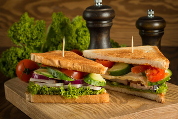 Close-up of two sandwiches with bacon, salami, prosciutto and fresh vegetables on rustic wooden cutting board. Club sandwich concept.