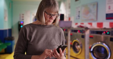 Serious senior woman using smartphone to read more of today's tragic world news standing at laundromat, Somber old lady reading distressing news alone at laundromat, 4k - Powered by Adobe