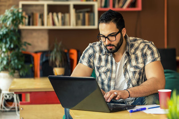Handsome bearded successful man with eyeglasses working with laptop in modern stylish coworking room with bookshelves, smart thinking