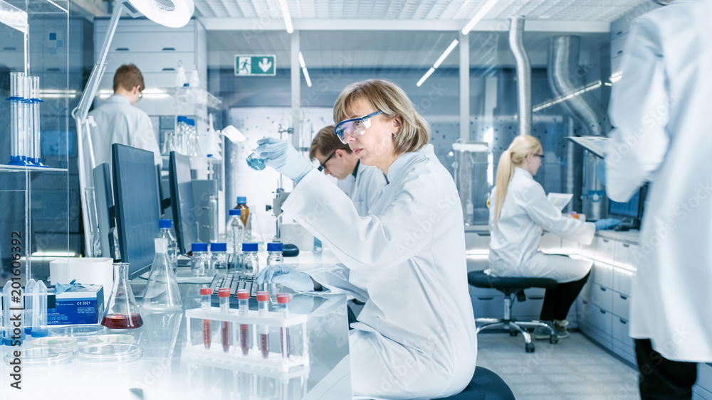 Wall mural female scientist analyzes liquid in the beaker and types down observations on her computer. she's wo