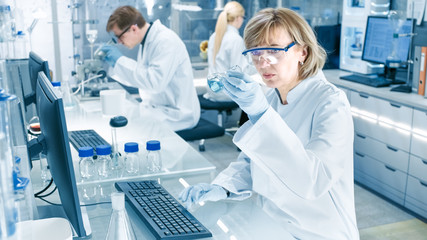 Female Scientist Analyzes Liquid in the Beaker and Types Down Observations on Her Computer. She's...