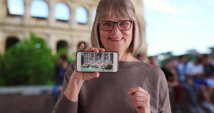 Joyful Caucasian old woman by Roman Colosseum showing video of Trevi Fountain, Smiling elderly female tourist in Rome showing off clip of Trevi Fountain on phone, 4k