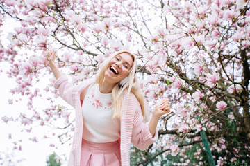 Cute blonde woman in pink clothes dancing in blooming gardens. Girl in headphones listens to music under blossom magnolia tree.