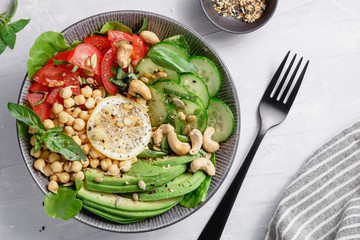 Top view of a healthy vegan lunch bowl with raw avocado, chickpeas, cashew, tomatoes, cucumber, greens and seeds over white kitchen table.