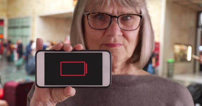 Old female at train station holding phone with low-battery indicator flashing, Caucasian female elder showing low-battery phone to camera at train station lobby, 4k