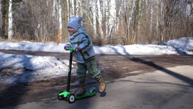 The child rides a scooter in the Park in early spring. Melting snow, snow and puddles on asphalt. Outdoor sports