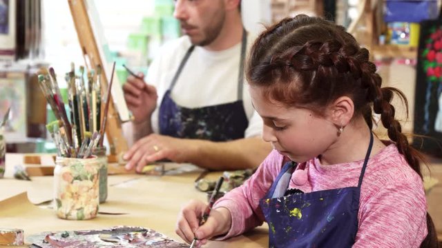 Little beautiful happy girl enjoying drawing at art studio smiling to the camera her father professional painter artist working on a painting on the background family creativity hobby artistry.