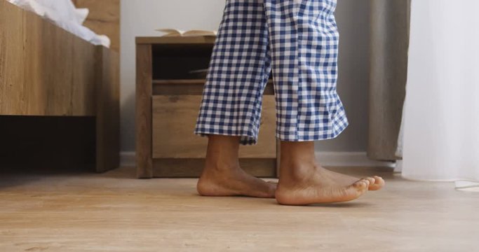 Close up of the female bare feet stepping on the floor from the bed in the morning and walking to the window. Indoors