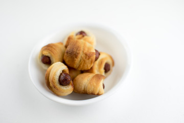 Home made pigs in a blanket. Sausages rolled in croissant dough baked and placed in a white serving dish. On white background.