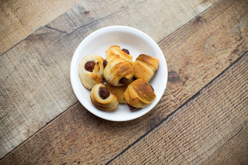 Home made pigs in a blanket. Sausages rolled in croissant dough baked and placed in a white serving dish. On wood background.