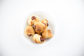 Home made pigs in a blanket. Sausages rolled in croissant dough baked and placed in a white serving dish. On white background.