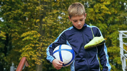Obraz premium Smiling teenage boy with a soccer ball in his hand and soccer boots on the shoulder against the background of the stadium. Sports training in the field.