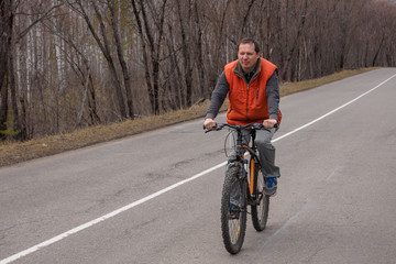 a man in an orange tank top rides on an asphalt road on a bike in the woods