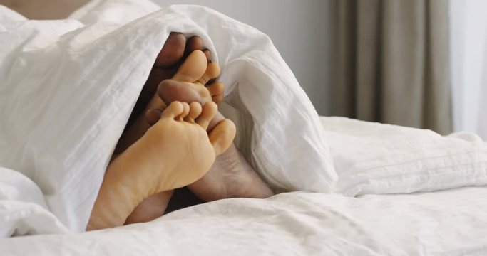 Close up of the African American male and female barefoot being seen from the white blanket in the bed in the moning. Inside