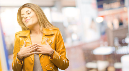 Beautiful young woman having charming smile holding hands on heart wanting to show love and sympathy at restaurant - obrazy, fototapety, plakaty