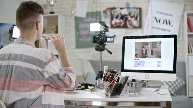 PAN of teenage boy showing eyeshadow palette to camera with led light standing on tripod while filming makeup tutorial