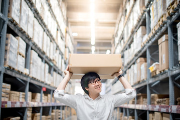 Young Asian man carry paper box over head between row of shelves in warehouse, shopping warehousing or working pick and packing concepts