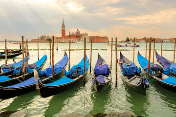 Fototapeta na wymiar Anchored gondolas standing in a row between piles against San Giorgio Madjore. Sunlight through the clouds. Venice