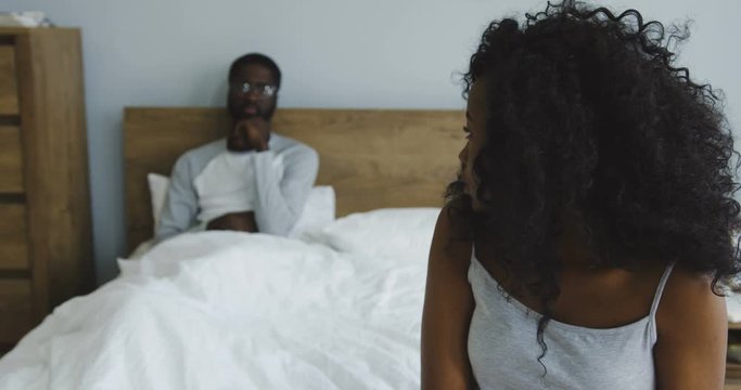 Sad young pretty African American woman in the pajama sitting on the corner of the bed, then turning her head and looking angrily at her husband or boyfriend who sitting in the bed behind. Inside