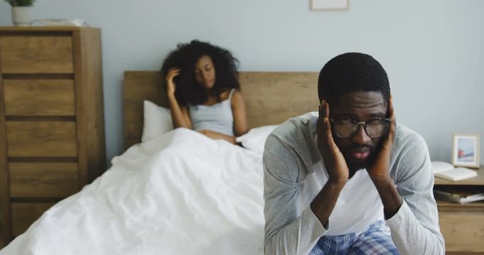 Sad young African American man in the pajama sitting on the corner of the bed, then turning his head and looking angrily at his wife or girlfriend who sitting in the bed behind. Indoor