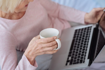 Senior gray haired woman holding the cup and using laptop.
