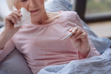 Sick elderly woman lying in a bed and holding the thermometer.
