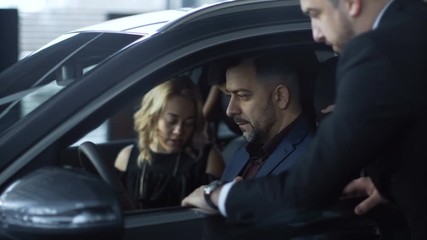 Side view of happy parents with kid sitting inside of car in auto shop and talking to consultant about purchase.