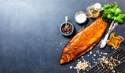 Smoked whitefish with salt and pepper in dish, fork and knife, lemon, basil, tomatoes, little cucumbers, parslay, grid for grilling, cherry wood chunks  on a black background, top view, overhead, flat