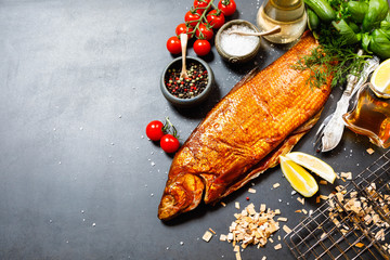 Smoked whitefish with salt and pepper in dish, fork and knife, lemon, basil, tomatoes, little cucumbers, parslay, grid for grilling, cherry wood chunks  on a black background, top view, overhead, flat