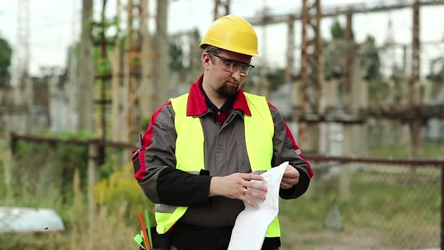 Worker with scheme of works on power station. Power man with construction documentation at power plant. Master builder in hard hat with design drawings
