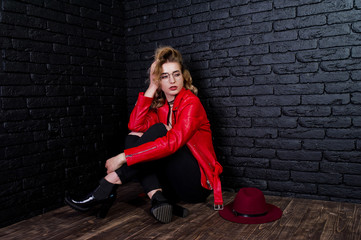 Fototapeta na wymiar Studio portrait of blonde girl with red hat, glasses and leather jacket posed against brick wall.