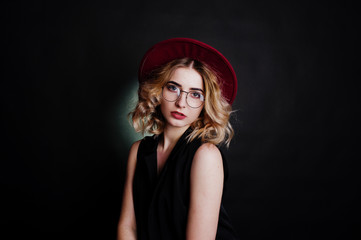 Studio portrait of blonde girl in black wear, red hat and glasses against dark background.