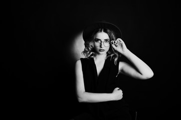 Studio portrait of blonde girl in black wear, red hat and glasses against dark background.