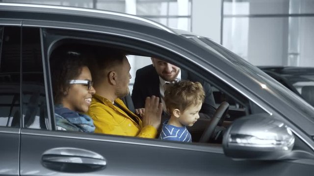 Side view of multiethnic couple with son sitting inside of car and talking to consultant testing new purchase,