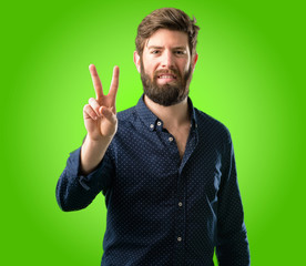 Fototapeta premium Young hipster man with big beard raising his fingers, number two over green background