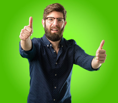 Young Hipster Man With Big Beard Stand Happy And Positive With Thumbs Up Approving With A Big Smile Expressing Okay Gesture Over Green Background