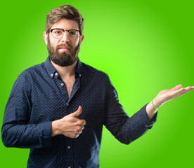 Young hipster man with big beard thumb up holding something in his empty hand and making ok gesture over green background