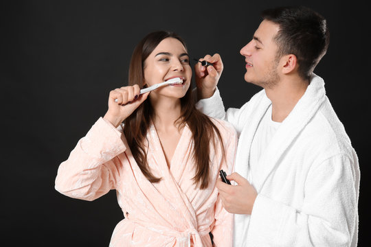 Young man applying mascara onto girlfriend's eyelashes while she brushing teeth on dark background
