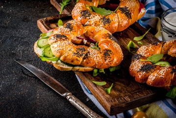 Variety of homemade bagels sandwiches with sesame and poppy seeds, cream cheese,  ham, radish, arugula, cherry tomatoes, cucumbers, on cutting board. dark concrete background copy space