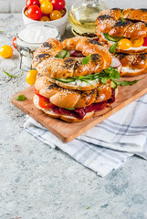 Variety of homemade bagels sandwiches with sesame and poppy seeds, cream cheese,  ham, radish, arugula, cherry tomatoes, cucumbers, white grey textured background copy space